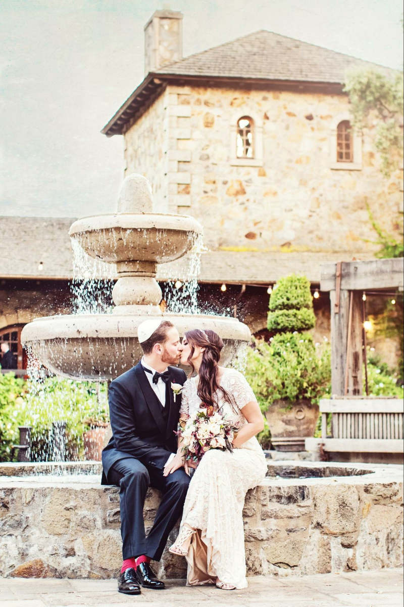 a bride and groom sitting on a stone wall in front of a fountain